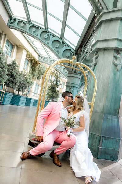 A couple in a pink suit and white dress kiss beside a gold luggage cart