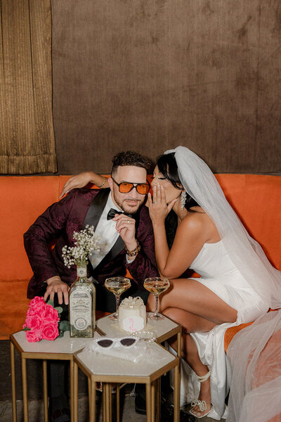 A bride leans in to whisper to her partner on an orange lounge booth